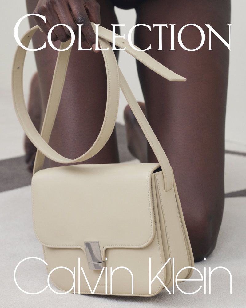 Cream-colored Calvin Klein purse with a silver closure, carried by a dark-skinned person with neutral manicure, kneeling indoors.
