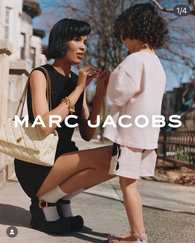 Fashion ad featuring an adult woman crouching next to a child outdoors, showcasing a designer handbag and apparel on a sunny day.