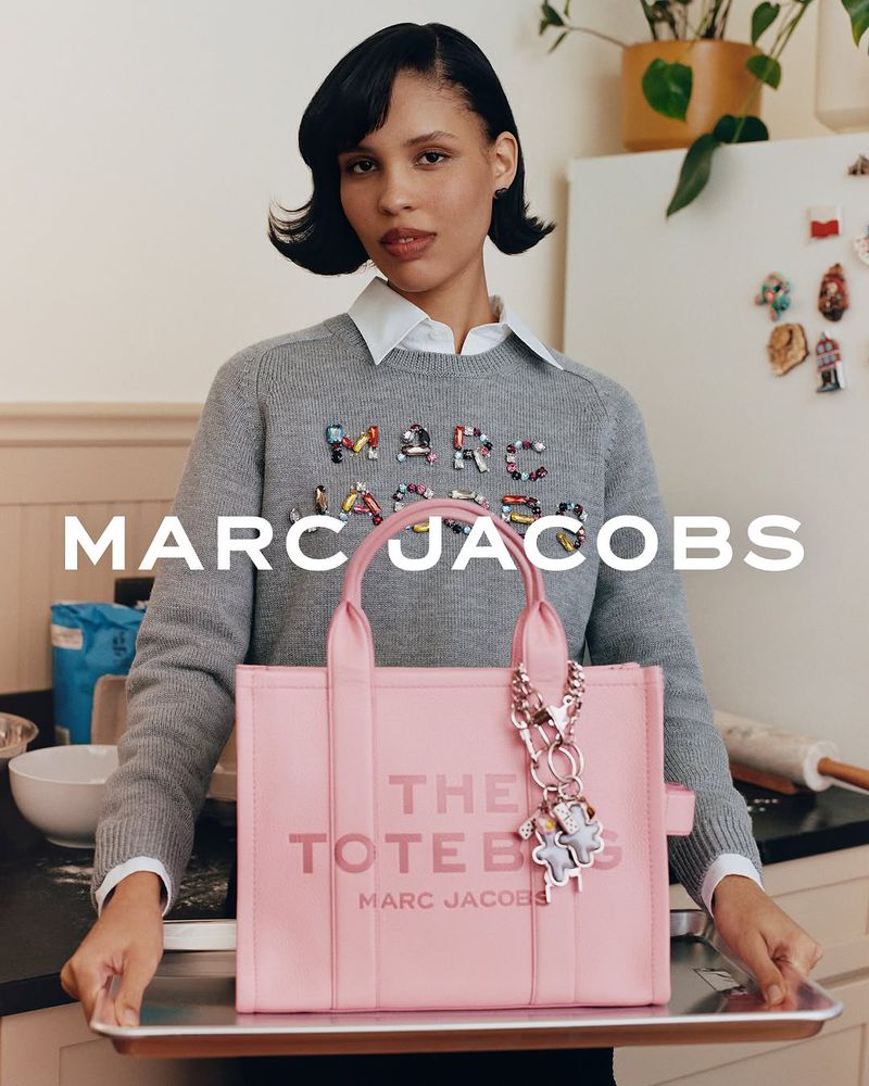 Adult female presents Marc Jacobs pink tote bag with charm in kitchen setting, wearing grey sweater. Fashion editorial lifestyle photography.