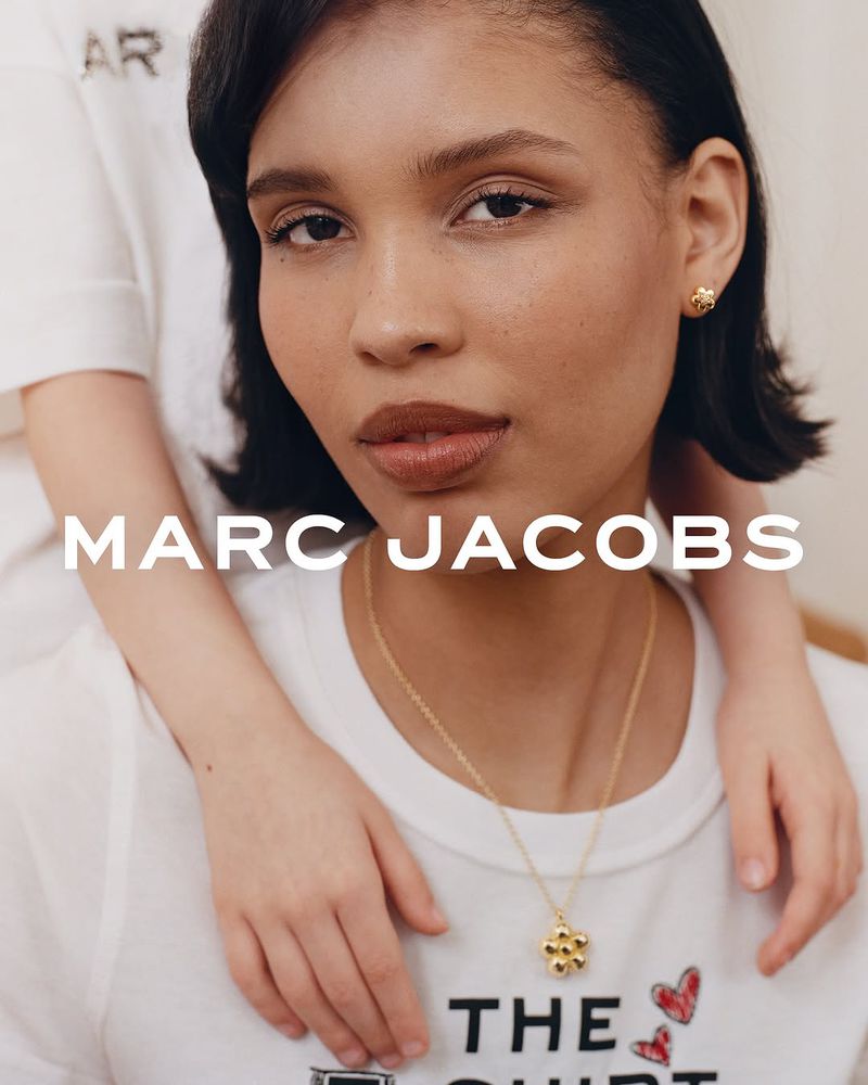 Close-up of an adult woman wearing jewelry, with another person behind her. The text 'MARC JACOBS' is prominently displayed.