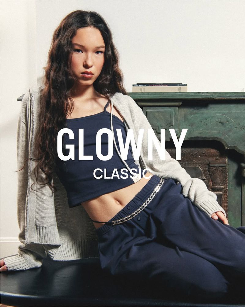 A young woman with long wavy brown hair poses in a navy crop top and sweatpants, wearing a grey zip up sweater, indoors, against a neutral background.