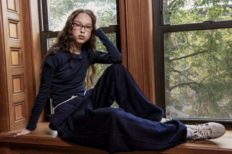 Young girl with long brown hair and glasses, wearing a navy outfit and Adidas sneakers, sits in a window with a green leafy background.