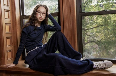 Model Kyla Malloy wearing navy blue loungewear and eyeglasses seated on a wooden window sill looking at the camera.