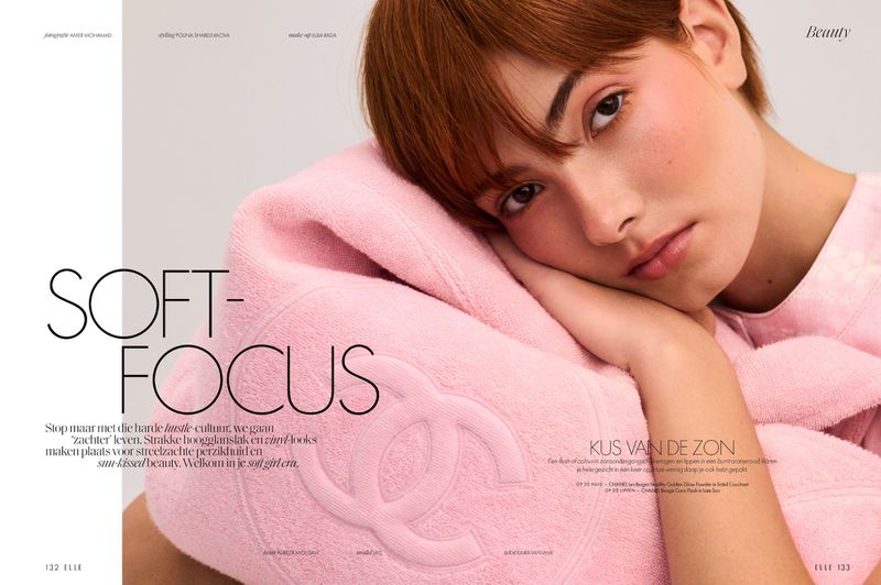 Close-up of a red-haired woman with light skin, resting her face on a light pink towel with a Chanel logo in an indoor setting.