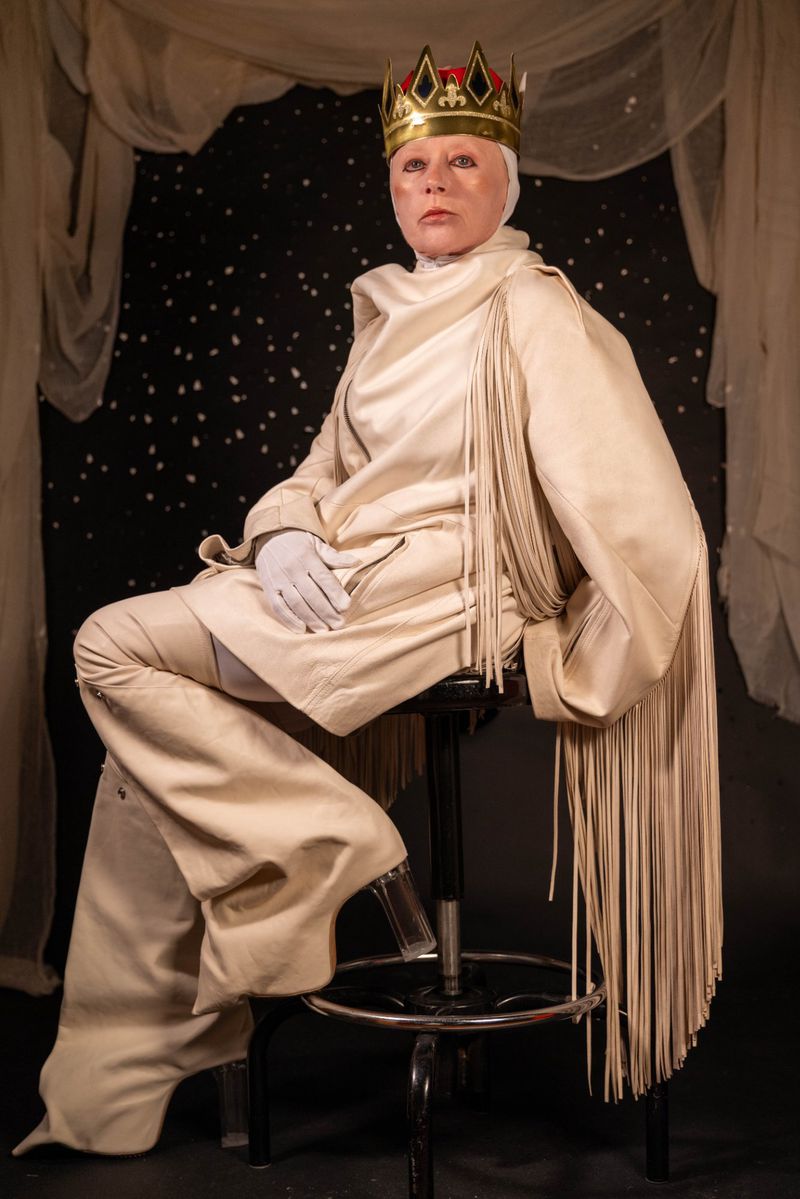Adult wearing a gold crown and a light-beige outfit with fringe, sitting on a stool in front of a dark star patterned backdrop.