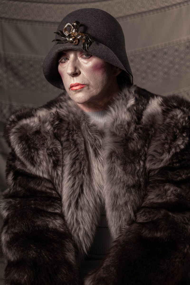Older woman with a grey hat and fur coat poses in a photo studio against a fabric backdrop. She has fair skin and red lipstick.