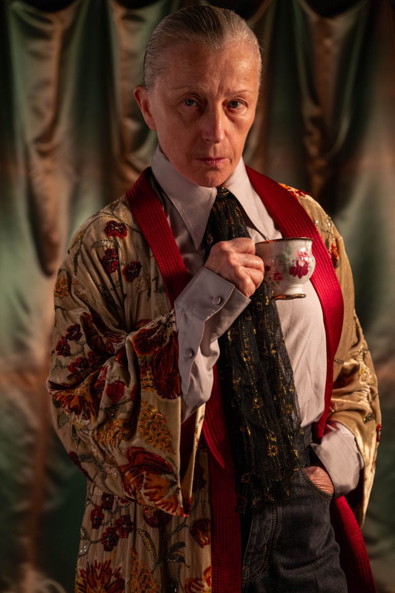 Elegant woman in a patterned robe holds a teacup against a draped fabric backdrop, showcasing a unique blend of sophistication and style.