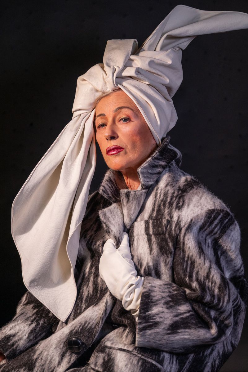 Elegant older woman with a cream-colored head wrap and a patterned black, gray and white coat poses against a dark gray backdrop.