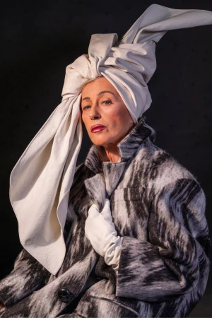 Elegant older woman with a cream-colored head wrap and a patterned black, gray and white coat poses against a dark gray backdrop.