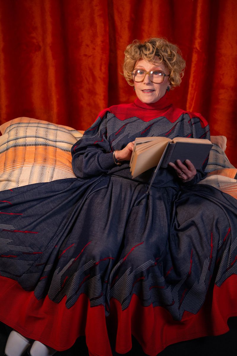 Mature woman with curly hair wearing glasses, reading a book while sitting on a bed in front of red curtains. Warm indoor lighting.