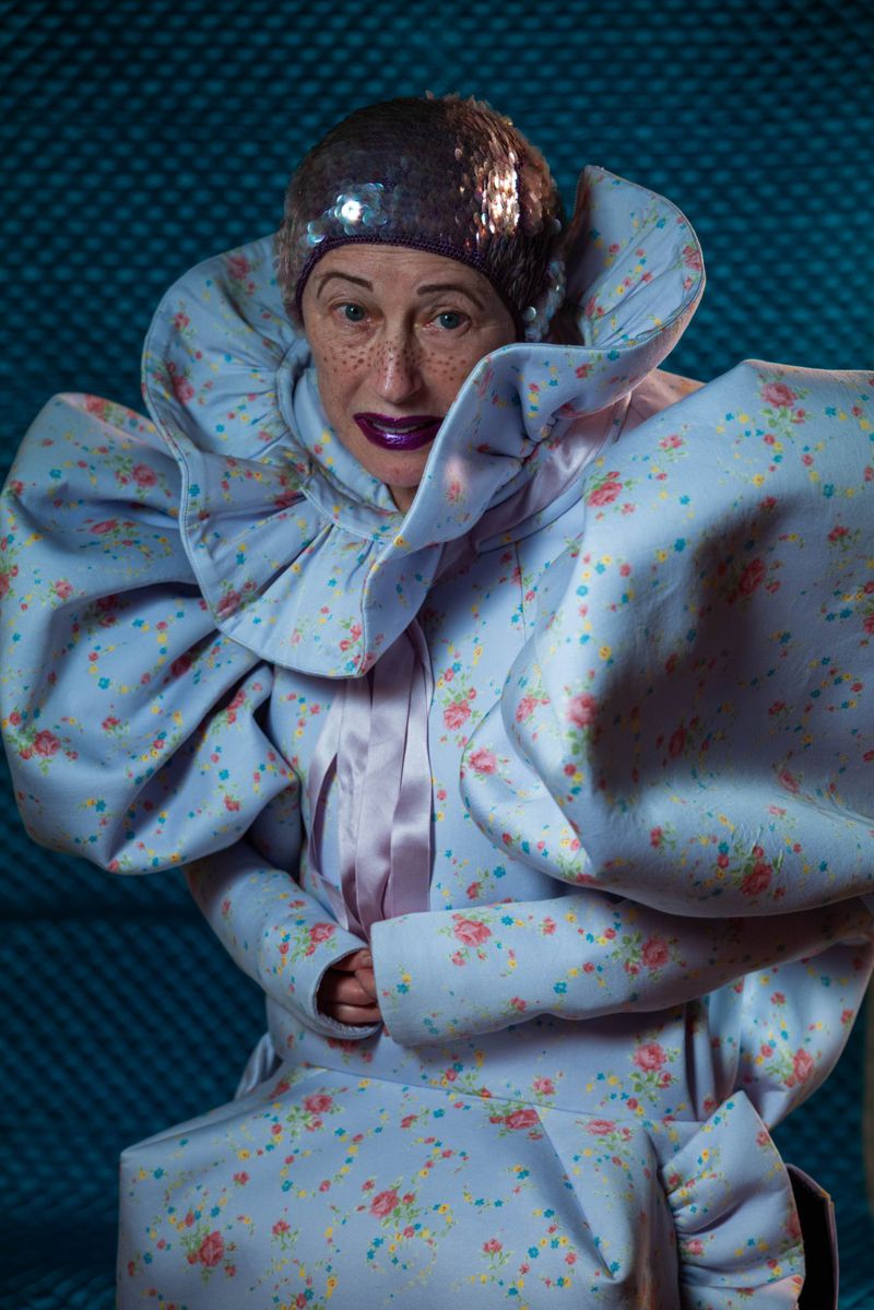 Portrait of an adult woman in a floral dress and sequined hat, looking at the camera, against a blue patterned background.