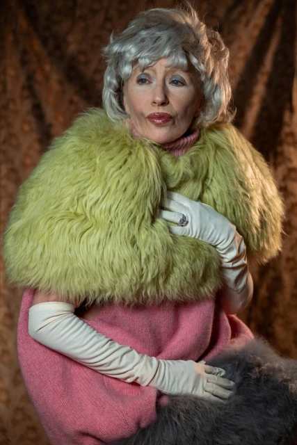 Mature adult female in photo studio wearing a bright green faux fur stole and pink sweater, paired with white opera gloves against brown backdrop.
