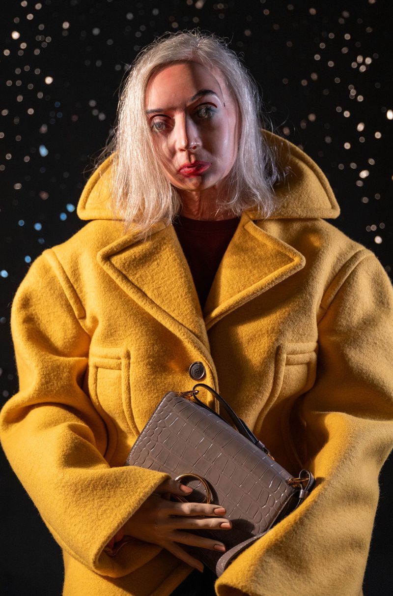 Woman in a yellow coat holds a grey purse against a star-field background, creating a striking fashion portrait.