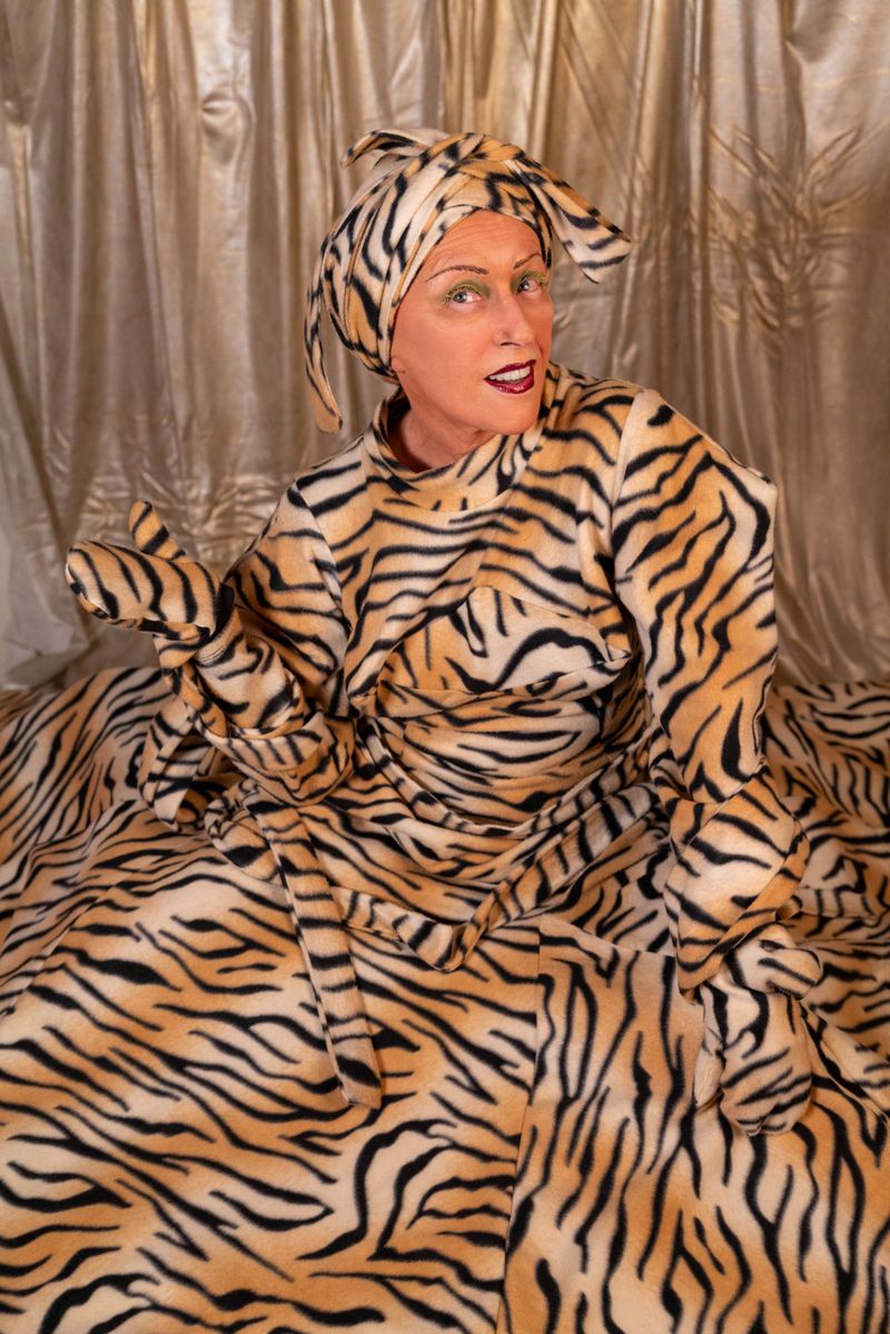 Person in tiger-striped outfit and head covering poses against gold curtain backdrop in a brightly lit, eye-level medium shot.