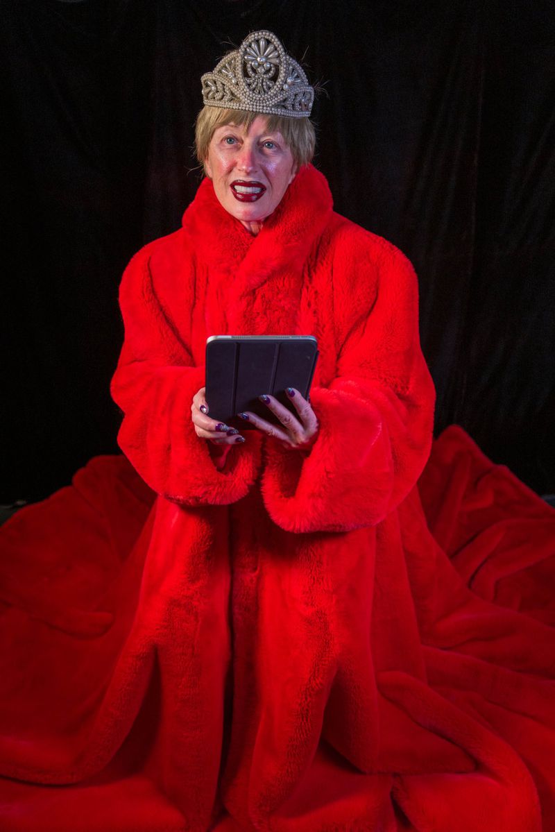 Woman in red faux fur coat and tiara holds a tablet against a black background, making a theatrical pose with open mouth.
