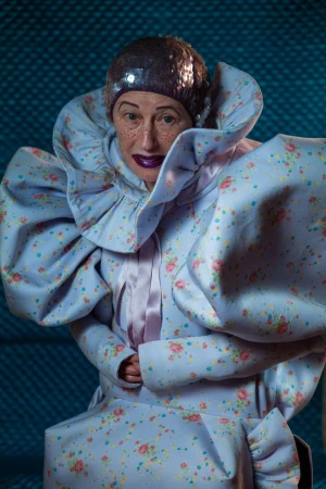 Cindy Sherman in an exaggerated floral print gown with a massive ruffled collar, voluminous puff sleeves, and a purple sequined cap.