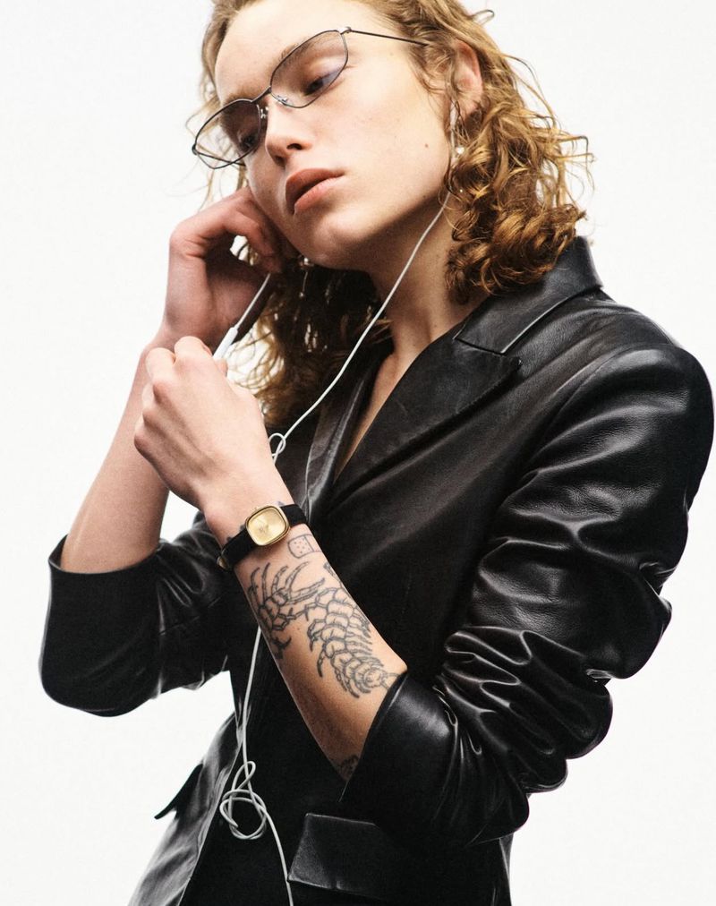 Model wearing a black leather jacket, wireframe glasses, and a gold watch while holding wired earphones against a white studio background.