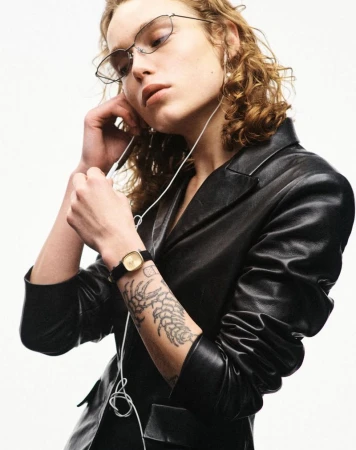 Model wearing a black leather jacket, wireframe glasses, and a gold watch while holding wired earphones against a white studio background.