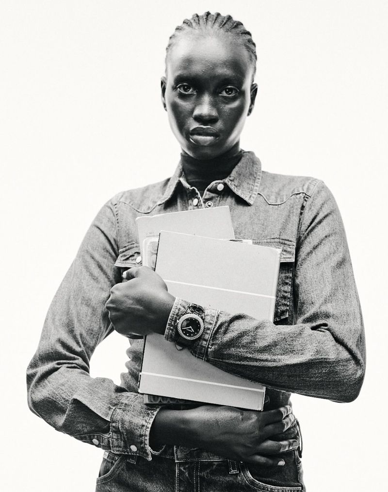 Monochrome portrait of a Black woman in a denim shirt, holding folders, set against a white background in a studio-style setting.