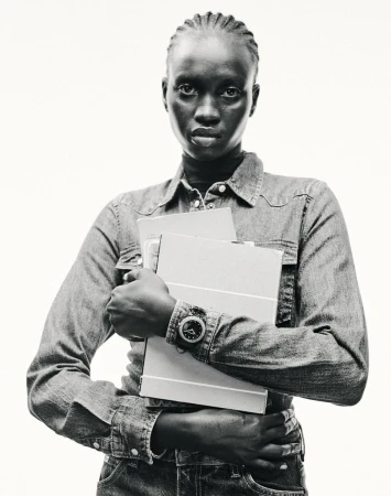 Monochrome portrait of a Black woman in a denim shirt, holding folders, set against a white background in a studio-style setting.