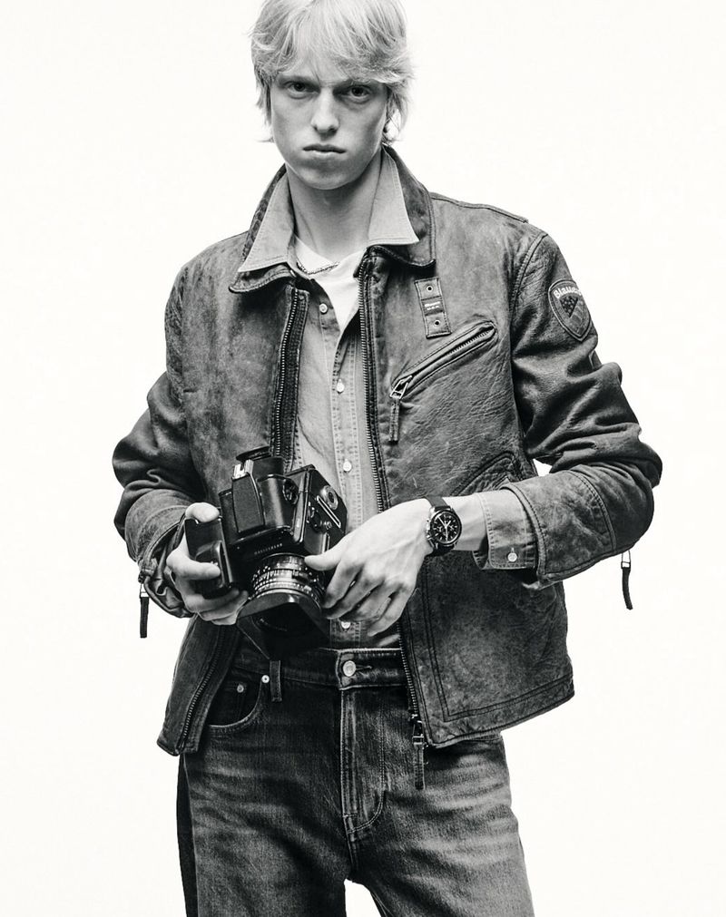 Monochrome portrait of a blond male holding a vintage camera, wearing a denim jacket and jeans against a plain white background.