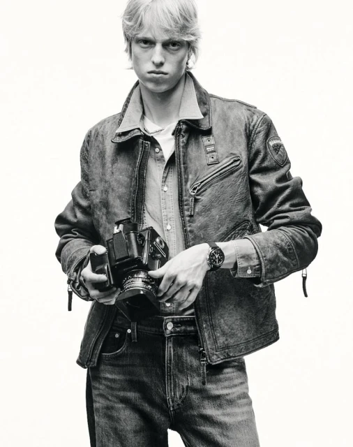 Monochrome portrait of a blond male holding a vintage camera, wearing a denim jacket and jeans against a plain white background.