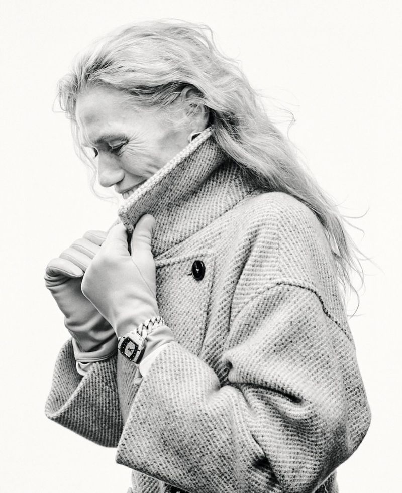 Elegant elderly woman in tweed coat and gloves looking down in black and white studio shot. Classic watch on wrist. Clean, bright background.