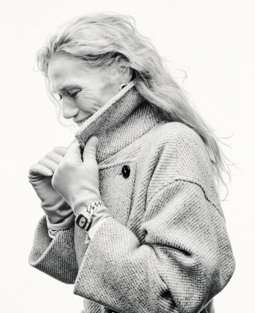 Elegant elderly woman in tweed coat and gloves looking down in black and white studio shot. Classic watch on wrist. Clean, bright background.
