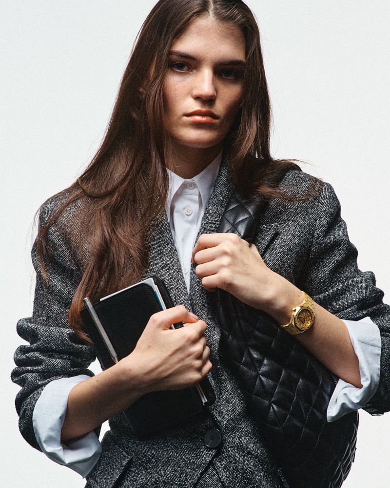 Portrait of a young woman with long hair holding a notebook, wearing a blazer and carrying a quilted leather shoulder bag on a white backdrop.