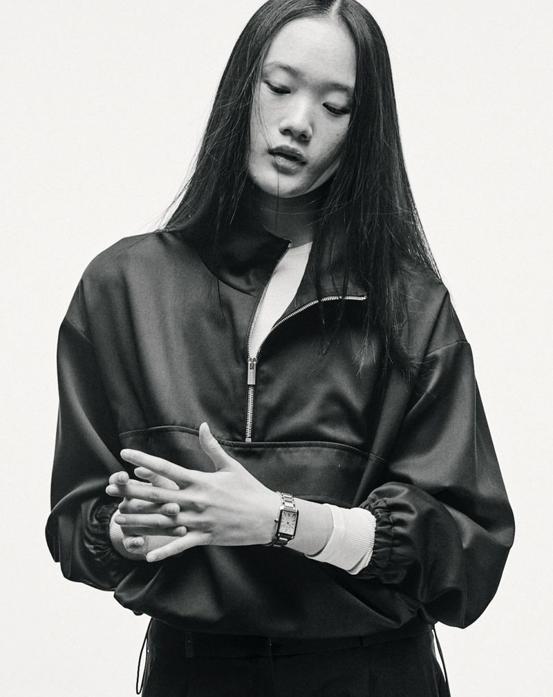 Black-and-white portrait of a young Asian woman with long hair wearing a quarter-zip jacket and a wristwatch, set against a light background.