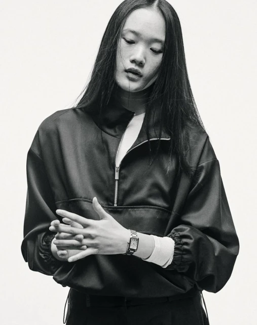 Black-and-white portrait of a young Asian woman with long hair wearing a quarter-zip jacket and a wristwatch, set against a light background.