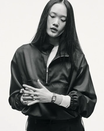Black-and-white portrait of a young Asian woman with long hair wearing a quarter-zip jacket and a wristwatch, set against a light background.