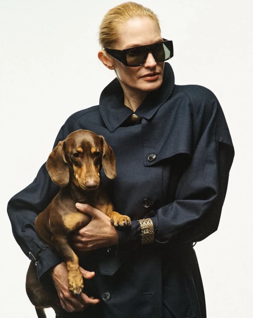 Adult woman wearing sunglasses and a navy coat holds a brown dachshund in a studio shot on a white backdrop.