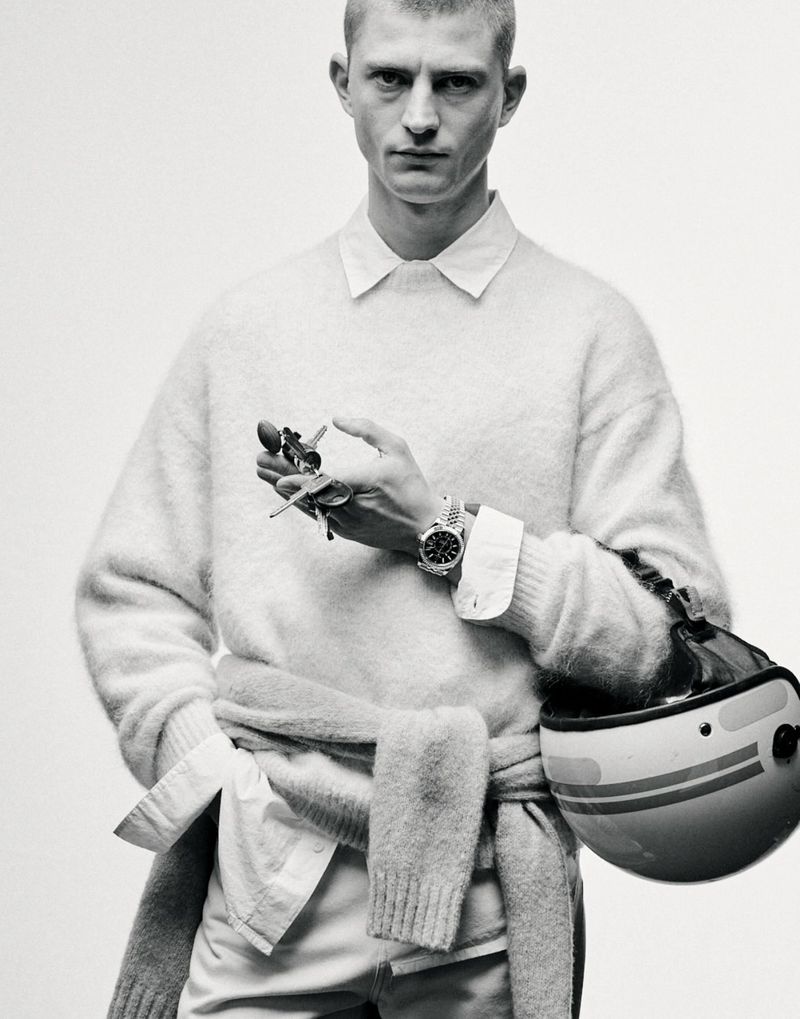 Monochrome portrait of a man in a sweater holding keys, with a motorcycle helmet nearby, showcasing a casual yet refined aesthetic.
