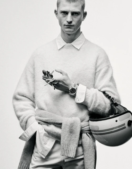 Monochrome portrait of a man in a sweater holding keys, with a motorcycle helmet nearby, showcasing a casual yet refined aesthetic.