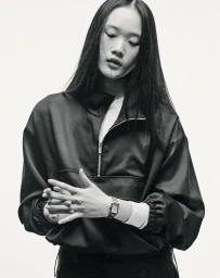 Black and white portrait of a woman with long dark hair wearing a black half-zip pullover and wristwatch against a white background.