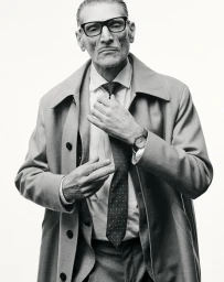 Black and white portrait of a mature man in thick glasses and an overcoat adjusting his polka dot necktie against a white background.
