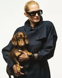 Woman wearing dark sunglasses and a navy blue trench coat holding a brown dachshund against a white studio background.