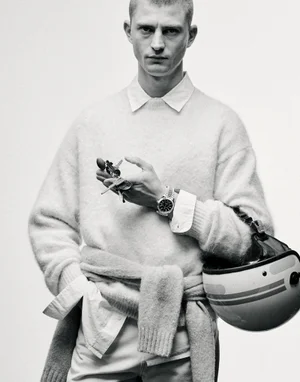 Model in a knit sweater and collared shirt holding keys and a motorcycle helmet against a white studio background.
