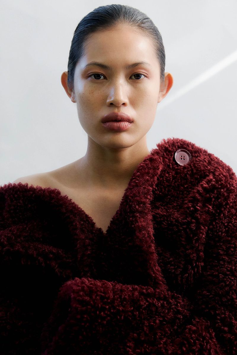 Portrait of a stylish adult woman with sleek black hair, wearing a maroon textured jacket with a button, posing in a studio.