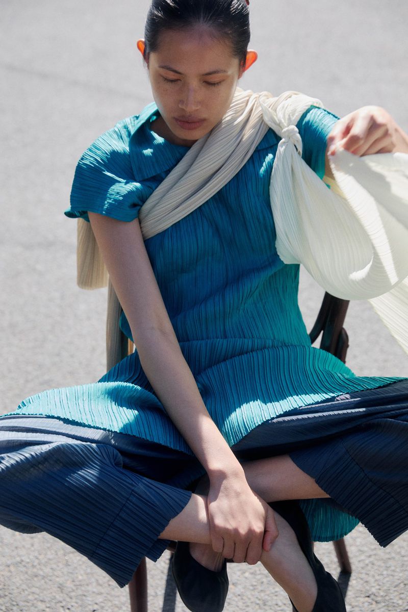 A woman with an Asian appearance sits on a chair wearing a teal dress with a cream shawl. She is outside on concrete.
