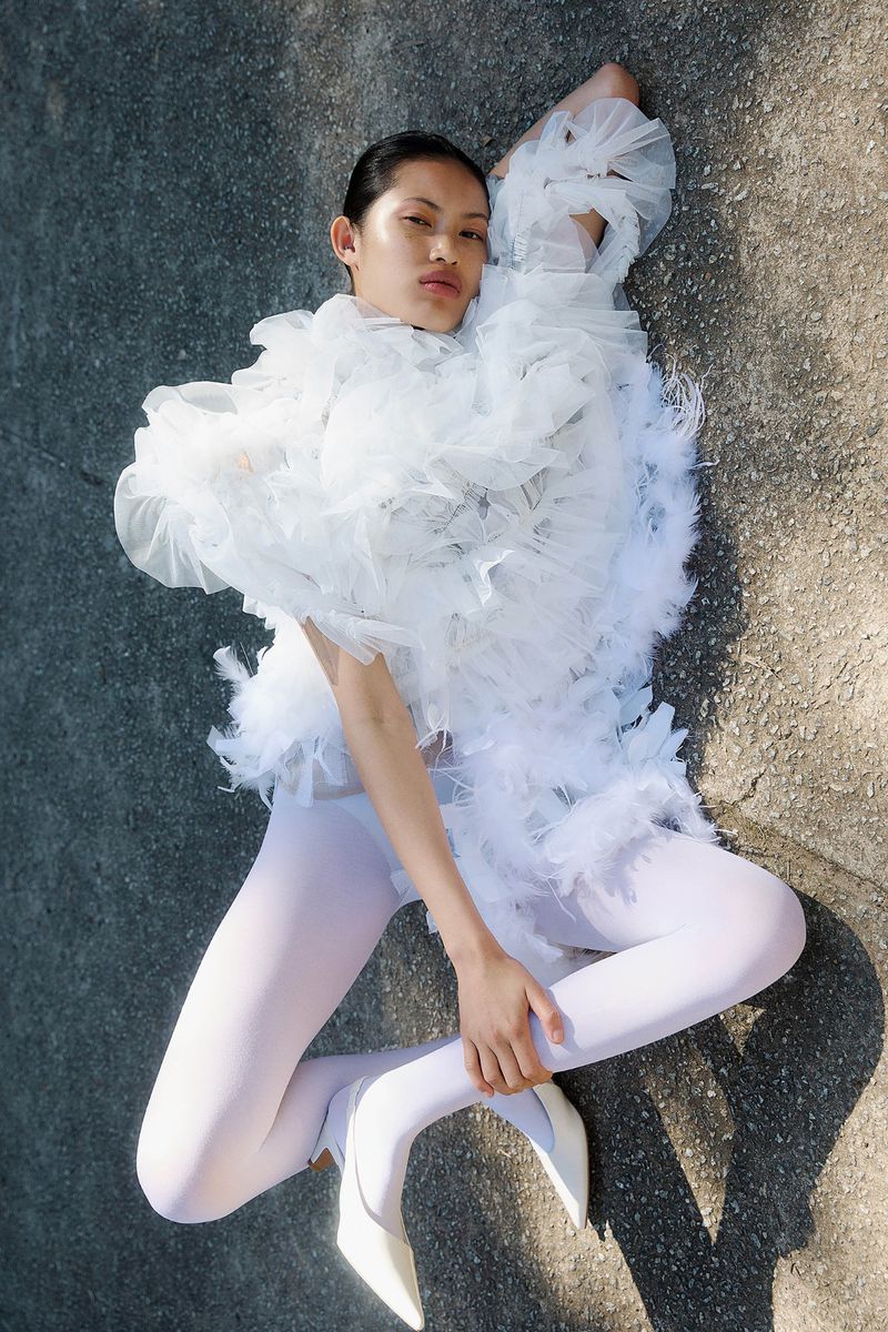 Fashion shot of a woman in a white ruffled garment lying on the ground with white tights and heels, shot from an eye-level angle.