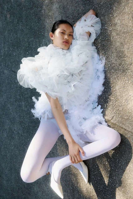 Fashion shot of a woman in a white ruffled garment lying on the ground with white tights and heels, shot from an eye-level angle.