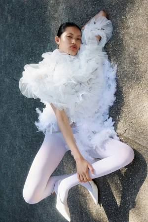 Fashion shot of a woman in a white ruffled garment lying on the ground with white tights and heels, shot from an eye-level angle.