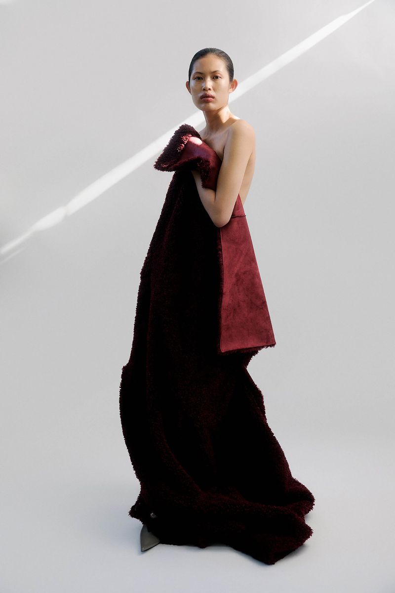 Elegant woman draped in a plush, burgundy fabric stands against a white background, captured in a fashion-forward, full-length portrait.
