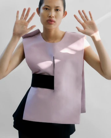 Woman poses in lilac top and black skirt against a white backdrop, raising her hands with fingers spread toward the camera.
