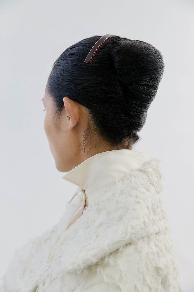 Elegant updo hairstyle on an adult female, featuring a hair comb accessory and white textured jacket, set against a plain white background.