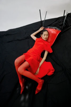 Fashion editorial shot featuring an Asian woman in a red dress and tights reclining on a red chair against a black and white backdrop.