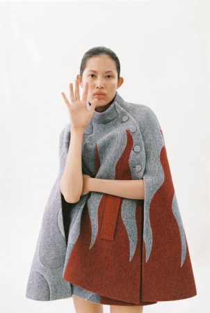 East Asian woman wearing a high-collar grey and rust wool cape posing with a raised hand against a plain white backdrop.
