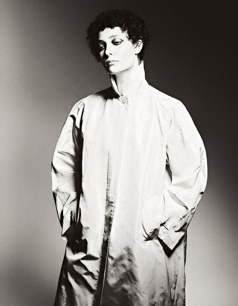 Monochrome portrait of a woman with short curly hair wearing a white, high-collared coat, set against a gradient grey background.